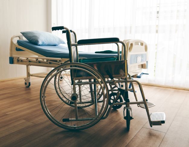 Wózek inwalidzki ustawiony obok łóżka rehabilitacyjnego w pomieszczeniu opieki medycznej.